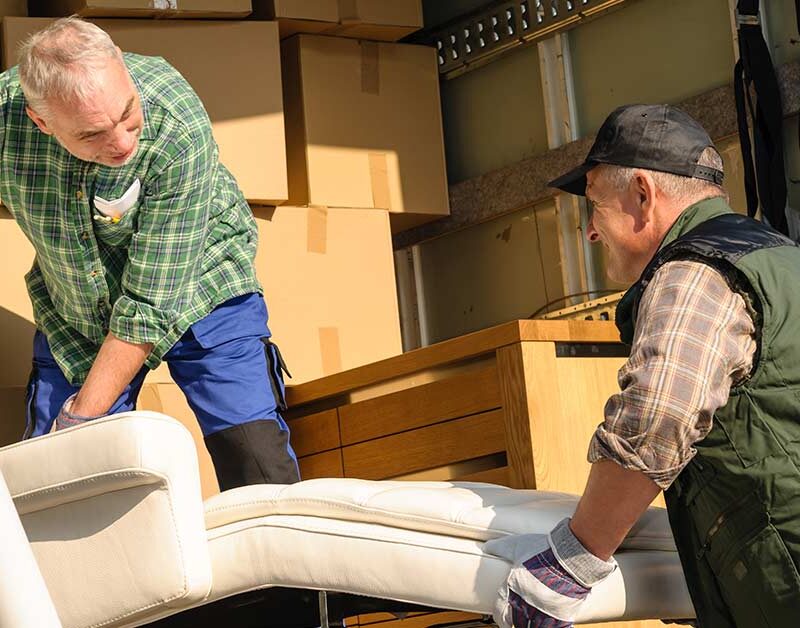 Zwei Männer heben in einem Umzugswagen voller Kisten ein Sofa an - Wohnen im Alter
